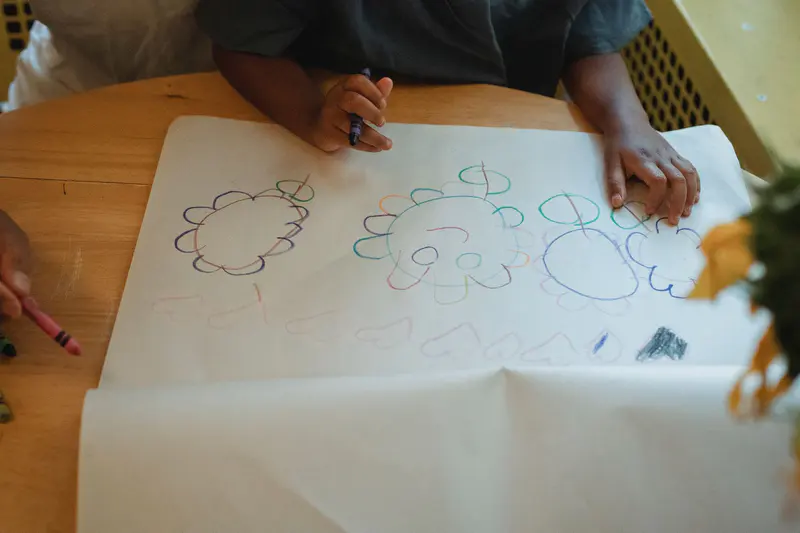 A child's hands drawing flowers in crayon on a large sheet of paper